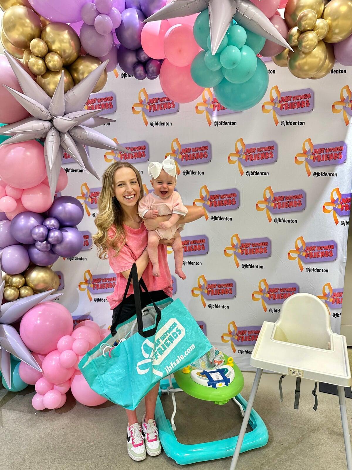 A mom with a large JBF shopping bag on her shoulder stands beside her husband who wears their toddler at a JBF sale.