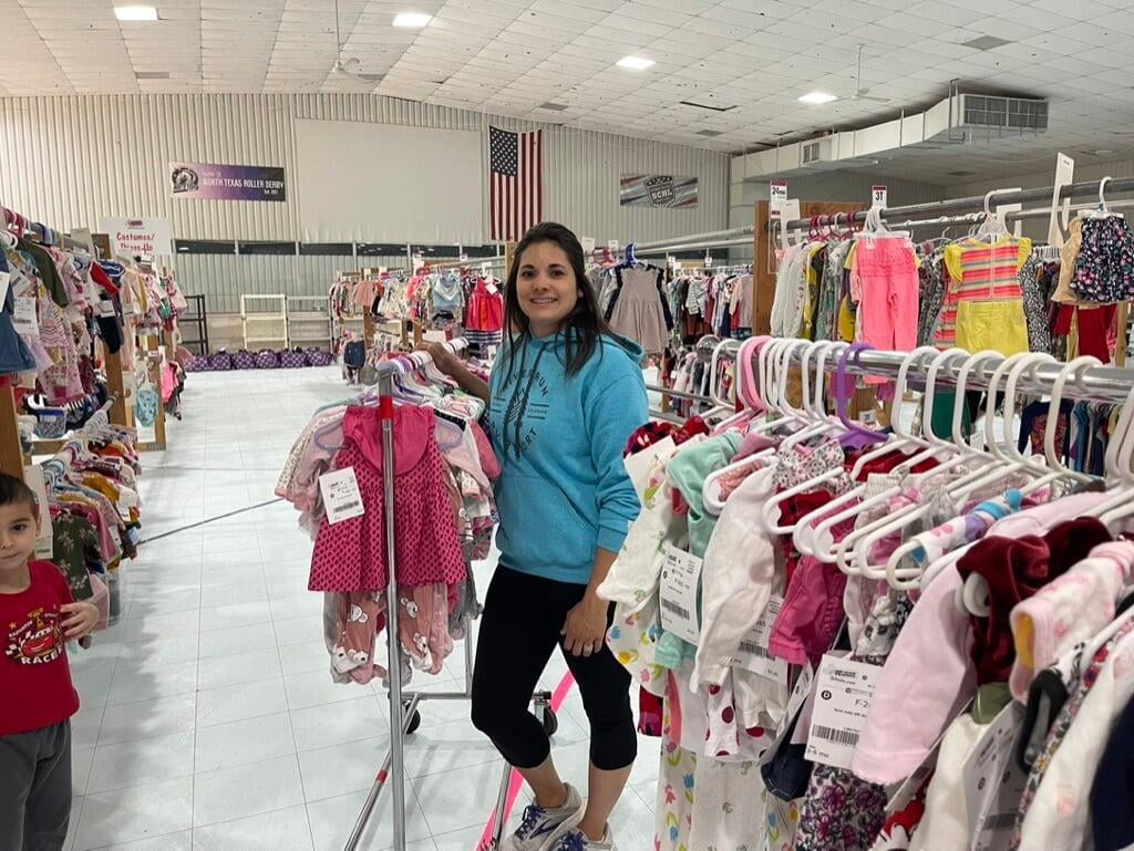 Mom wears baby and holds toddler boy clothes in each hand. Second mom shops behind her from the racks of boys clothes.